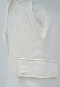 Jogging en coton blanc avec poches latérales et une poche à rabat avec une étiquette. Texture lisse avec taille à cordon. Design minimaliste.