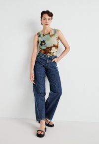 Top senza maniche multicolore con motivo tie-dye verde, marrone e crema, abbinato a jeans in denim blu a vita alta e sandali neri con plateau.