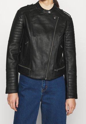 Frau trägt eine schwarze gesteppte Leder-Bikerjacke mit asymmetrischem Reißverschluss und blaue Jeans, auf Taillenhöhe gekürzt.