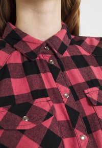 Red and black checkered flannel shirt with a pointed collar, silver snap buttons, and chest pockets. Fabric texture is soft and thick.
