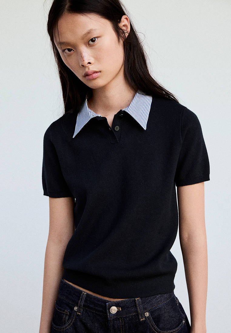 Jeune femme portant un pull noir à manches courtes avec un col rayé bleu et blanc, debout devant un fond clair uni.