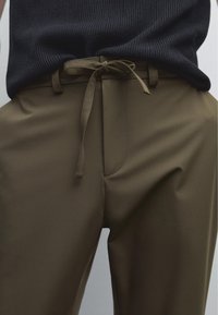 Pantalon vert olive avec une texture lisse, doté d'une poche avant, d'une taille avec un ruban noué et d'une coupe décontractée.