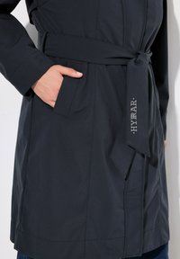 Cappotto impermeabile nero con vita regolabile, tasche laterali e un dettaglio di branding discreto. Texture liscia con una vestibilità su misura.