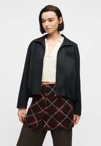 Γυναίκα με κοντά σκούρα μαλλιά που φοράει μαύρο μπουφάν με φερμουάρ πάνω από λευκή πουλόβερ με V-λαιμόκοψη και καρό καφέ φούστα με μοτίβο διαμαντιού.