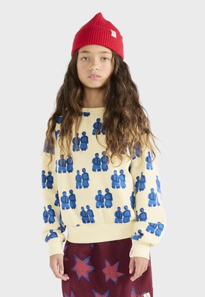 Cream-colored sweatshirt featuring a pattern of blue figures, paired with a maroon skirt adorned with red stars. The model wears a red knit beanie.