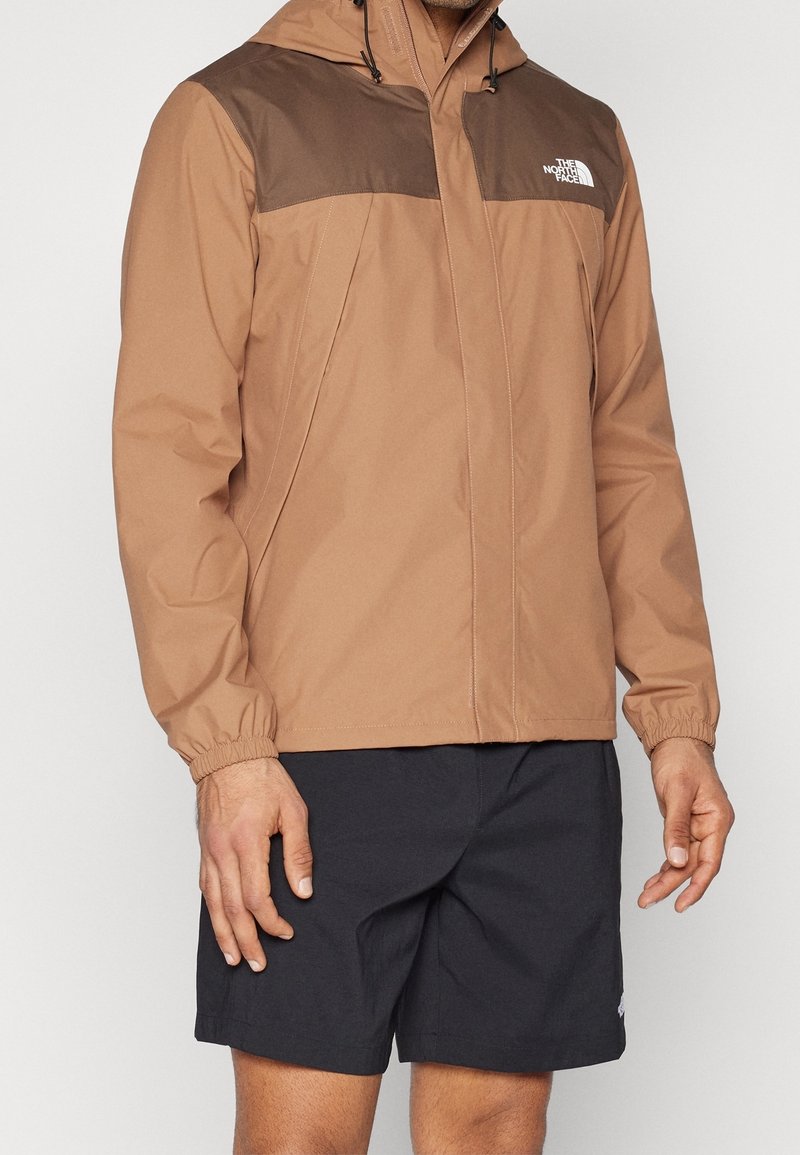 Veste imperméable marron avec une zone d'épaule marron foncé contrastante, des poignets élastiques, une fermeture éclair intégrale et une capuche. Portée avec un short foncé.