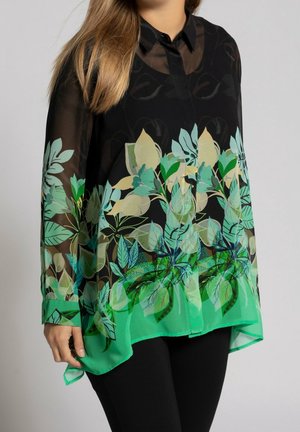 Femme portant un chemisier noir transparent avec des motifs floraux feuillus verts, beiges et bleus et un pantalon noir uni sur un fond uni.
