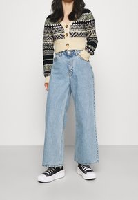 Gestrickte Strickjacke in Creme und Marineblau mit geometrischen Mustern, kombiniert mit hoch taillierten, hellblauen Jeans mit weitem Bein und schwarzen Turnschuhen.