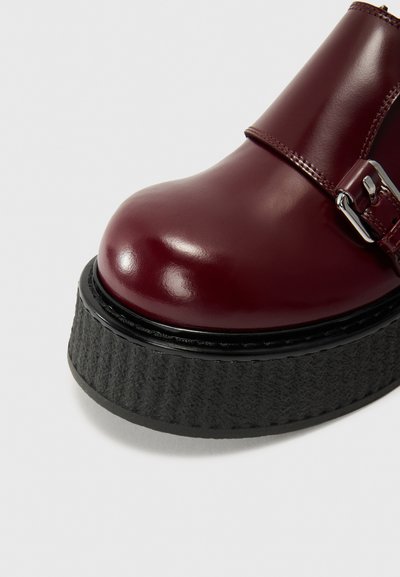 Μπορντό παπούτσι με τακούνι από πατέντα δέρμα, με λεία επιφάνεια, που διαθέτει υφή μαύρης βάσης και μεταλλική αγκράφα ως διακόσμηση.