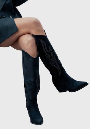 Jambes croisées portant des bottes hauteur genou en suède noir avec coutures décoratives et bouts pointus, associées à une jupe noire.