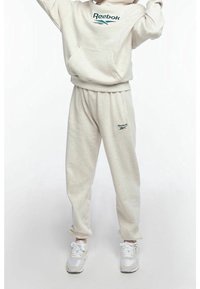 Osoba oblečená v béžové mikině Reebok a odpovídajících teplácích s bílými teniskami, stojící na čistém světlém pozadí.