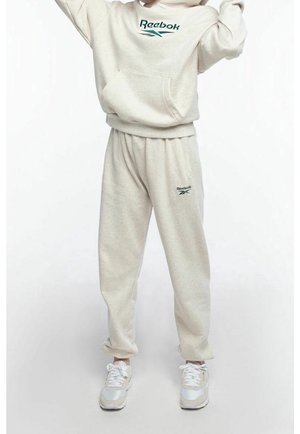 Person iført beige Reebok hoodie og matchende træningsbukser med hvide sneakers, stående mod en ensfarvet lys baggrund.