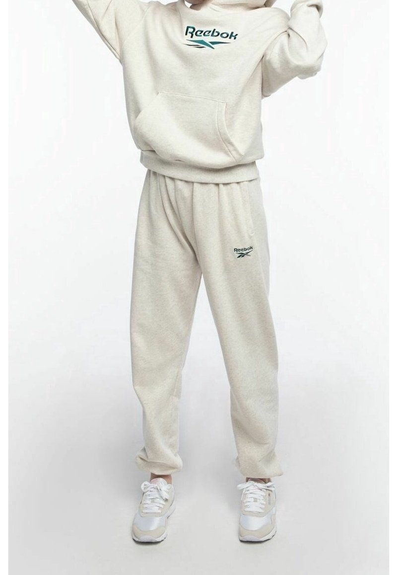 Osoba oblečená v béžové mikině Reebok a odpovídajících teplácích s bílými teniskami, stojící na čistém světlém pozadí.