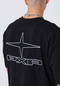 Camiseta negra de manga corta con un estrellita bordada blanca y el texto "PXP" en la parte de atrás. Tela de algodón suave con un ajuste relajado.