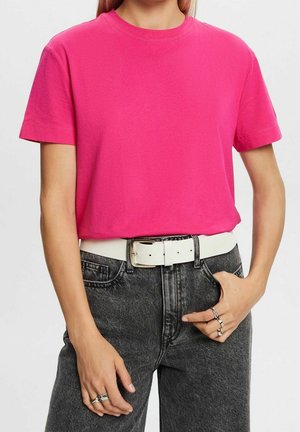 Persoon draagt een felroze kort mouwen shirt, ingestopt in zwarte gewassen jeans met een hoge taille, met een witte riem en zilveren ringen aan de vingers.