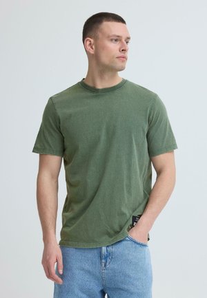 Nuori mies, jolla on vihreä lyhythihainen t-paita ja vaaleansiniset farkut, seisoo yhdellä kädellä taskussa ja katsoo sivulle.