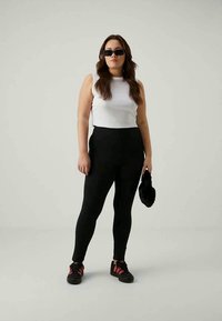 Mulher com óculos de sol pretos, blusa branca sem mangas, leggings pretas e sapatilhas pretas com detalhes em rosa, segurando uma pequena bolsa preta.