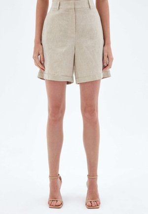 Short beige taille haute sur mesure avec revers, porté avec des sandales à talons hauts nude à brides la cheville, sur un mannequin debout.