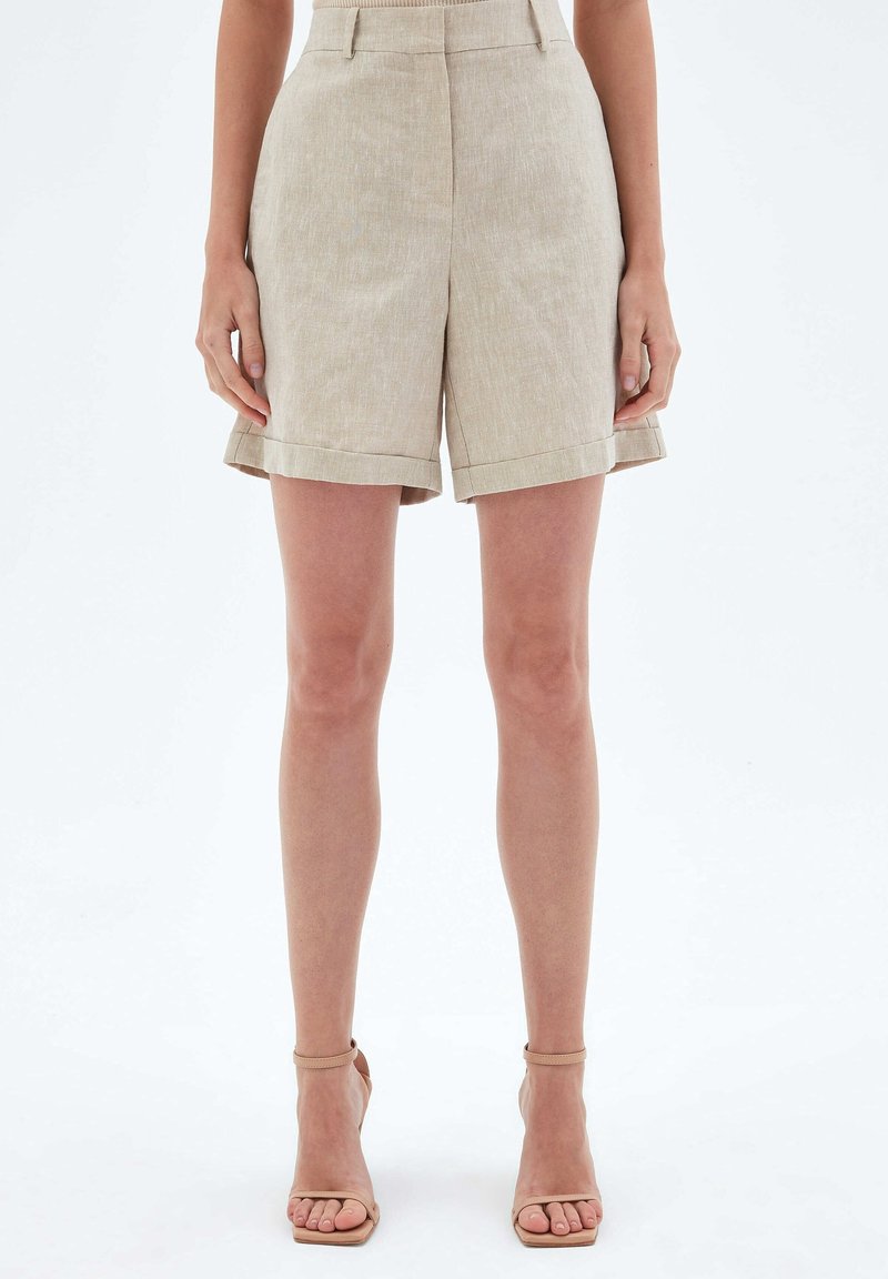 Short beige taille haute sur mesure avec revers, porté avec des sandales à talons hauts nude à brides la cheville, sur un mannequin debout.