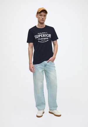 Marineblå grafisk t-skjorte med hvit tekst, lyseblå jeans i denim og hvite joggesko. Modellen har på seg en tanfarget cap; minimalistisk stil.