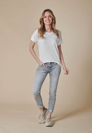 Frau steht lächelnd da, trägt ein weißes T-Shirt, hellgraue hochgekrempelte Jeans und beige Sneakers vor beigem Hintergrund.