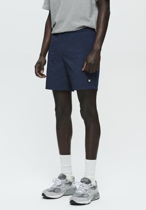 Shorts bleu marine avec une ceinture élastique, des poches latérales et un petit logo blanc ; associés à des chaussures de sport grises et des chaussettes blanches à mi-mollet.