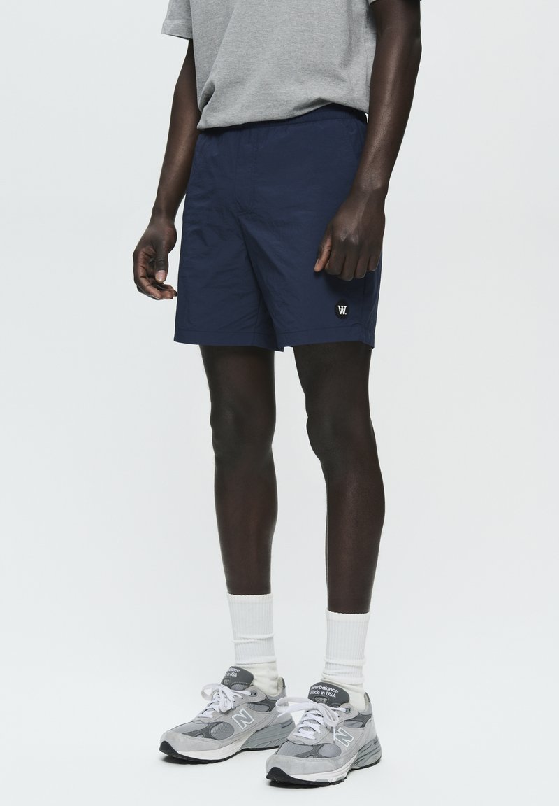 Pantaloni corti blu navy con una cintura elastica, tasche laterali e un piccolo logo bianco; abbinati a scarpe da ginnastica grigie e calze bianche. 