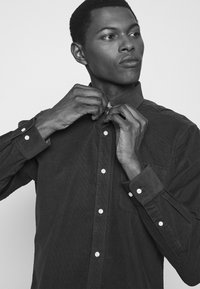 Overhemd met knopen van donkere stof, voorzien van witte knopen, een borstzak en een gestructureerde kraag. Subtiele textuur zichtbaar.