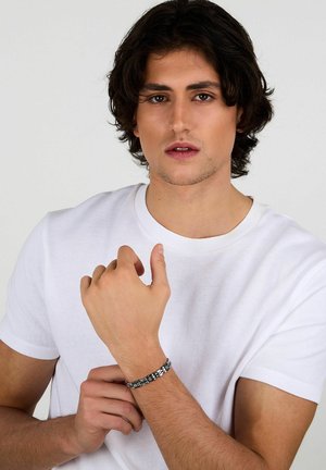 Giovane uomo con capelli scuri che indossa una camicia bianca aggiusta un bracciale a catena argento e nero sul suo polso davanti a uno sfondo semplice.