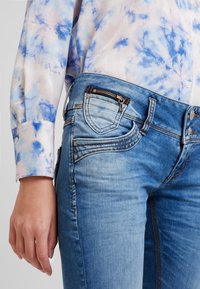 Světlé modré džíny z denimu se zipovou kapsou a podrobným prošíváním, v kombinaci s blůzkou s květinovým vzorem v pastelových barvách. Detailní záběr na oblast pasu.