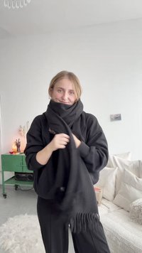 Foulard nero, tessuto morbido, bordi frangiati, avvolto intorno al collo. Maglione e pantaloni neri indossati. Stanza chiara con tessuti morbidi sullo sfondo.