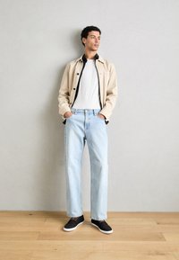 Chaqueta beige claro sobre una camisa negra, camiseta blanca y jeans rectos azul claro. Zapatillas negras con suelas blancas. Fondo de suelo de madera.
