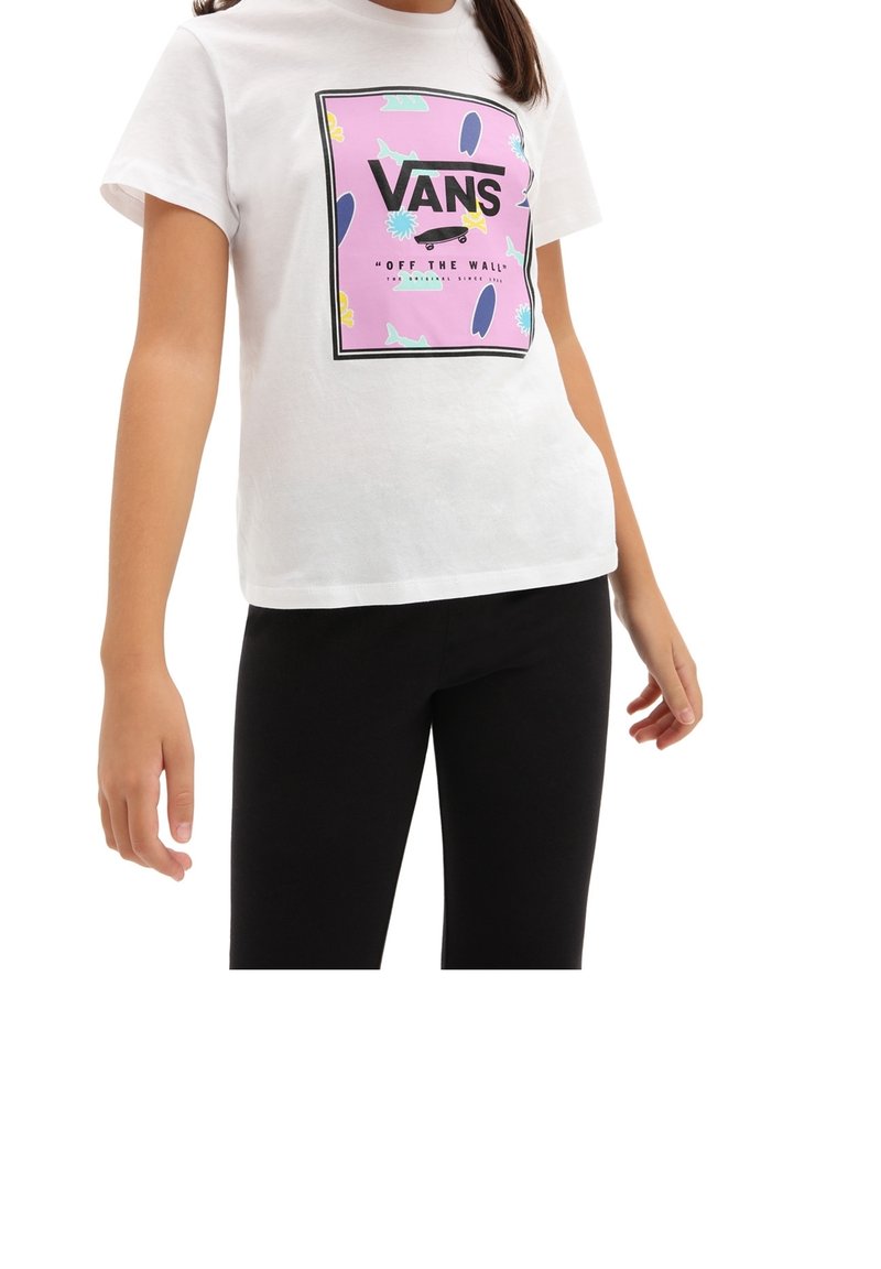 Personne portant un t-shirt blanc Vans avec un motif coloré de skateboard et un pantalon noir, debout les bras légèrement écartés.
