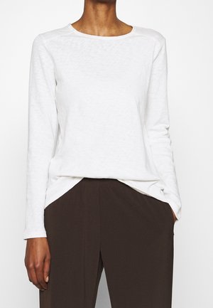 Persoon draagt een eenvoudige witte lange mouwen blouse, ingestopt in een hoog getailleerde donkerbruine broek, met één hand in de zak.