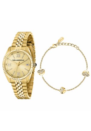 Montre en or avec chiffres romains et fenêtre date à côté d'un délicat bracelet chaîne en or orné de trois pendentifs en forme de cœur sertis de cristaux et d'un fermoir mousqueton.