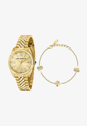 Guldkurv med romertal og datovindue ved siden af delikat guldkædearmbånd med tre hjertevedhæng pyntet med krystaller og hummerlås.