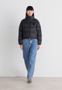 Donna che indossa un giubbotto piumino nero con il logo Calvin Klein, jeans azzurri chiari e tacchi aperti neri, mentre cammina in avanti.