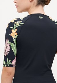 Čierny rash guard s kvetinovým dizajnom na rukávoch. Má krátke rukávy, ploché švy a malé logo na zadnej strane. Hladká textúra materiálu.