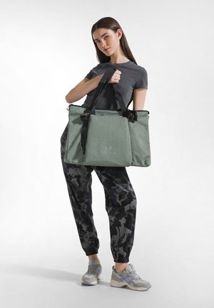 Zelená taška přes rameno z texturované látky s dvojitými horními madly, černými detaily a logem. Model má na sobě vzorované tepláky a tenisky.