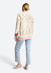 Strickjacke in Creme mit einem strukturierten Muster, versehen mit braunen Sprenkeln, ribbeligem rosafarbenem Saum und kontrastfarbigen Bündchen. Kombiniert mit hellen Denim-Hosen.