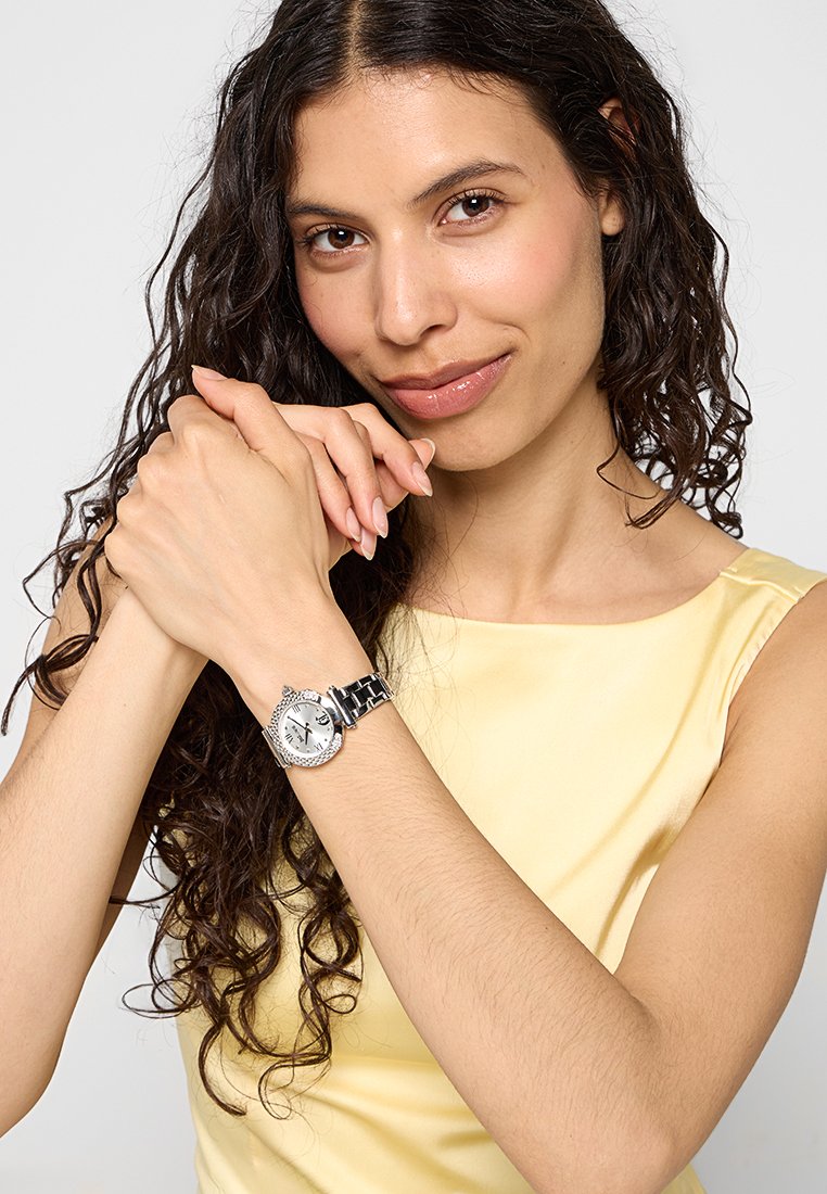 Orologio da polso in argento con quadrante bianco rotondo, bracciale in argento e accessori decorativi. Il modello indossa un top in satin giallo.