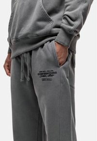 Grey cotton hoodie with a front pocket and ribbed cuffs, paired with matching sweatpants featuring embroidered branding and drawstrings.