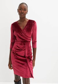 Abito in velluto cremisi con maniche lunghe, scollatura a V e dettagli arricciati. Presenta un design avvolgente laterale e sottili accenti di glitter. Tessuto morbido.