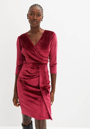 Robe en velours cramoisi à manches longues, décolleté en V et détails froncés. Présente un design drapé sur le côté et de subtils accents pailletés. Texture lisse.