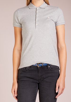 Kobieta ubrana w jasnoszarą koszulkę polo z krótkim rękawem, ozdobioną guzikami i małym haftowanym logo, w połączeniu z czarnymi dżinsami i niebieskim paskiem.