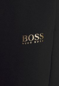 Černá látka s hladkým povrchem zdobena zlatým kovovým logem s nápisem "BOSS HUGO BOSS." Design je minimalistický a moderní.