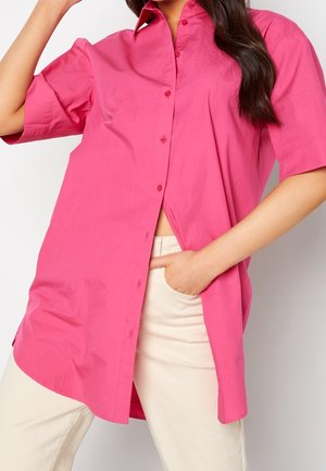 Mujer con una camisa abotonada de gran tamaño de color rosa brillante con mangas cortas, combinada con pantalones beige claro, mano en el bolsillo.