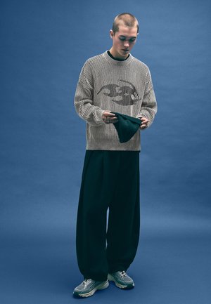 Striped knit sweater in beige with a graphic print, loose dark green trousers, and grey sneakers. Background is solid blue.