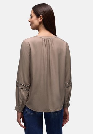 Kvinde med langt brunt hår iført en taupe-bluse med detaljeret indsamling på ryggen og mønstrede bånddekorationer på ærmerne, kombineret med blå jeans.