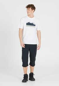 Valkoinen T-paita, jossa on vuorikuva ja "ULKOILMAHARRASTAJA" -teksti, yhdistettynä mustiin cropattuihin housuihin ja tummiin vaelluskengiin.